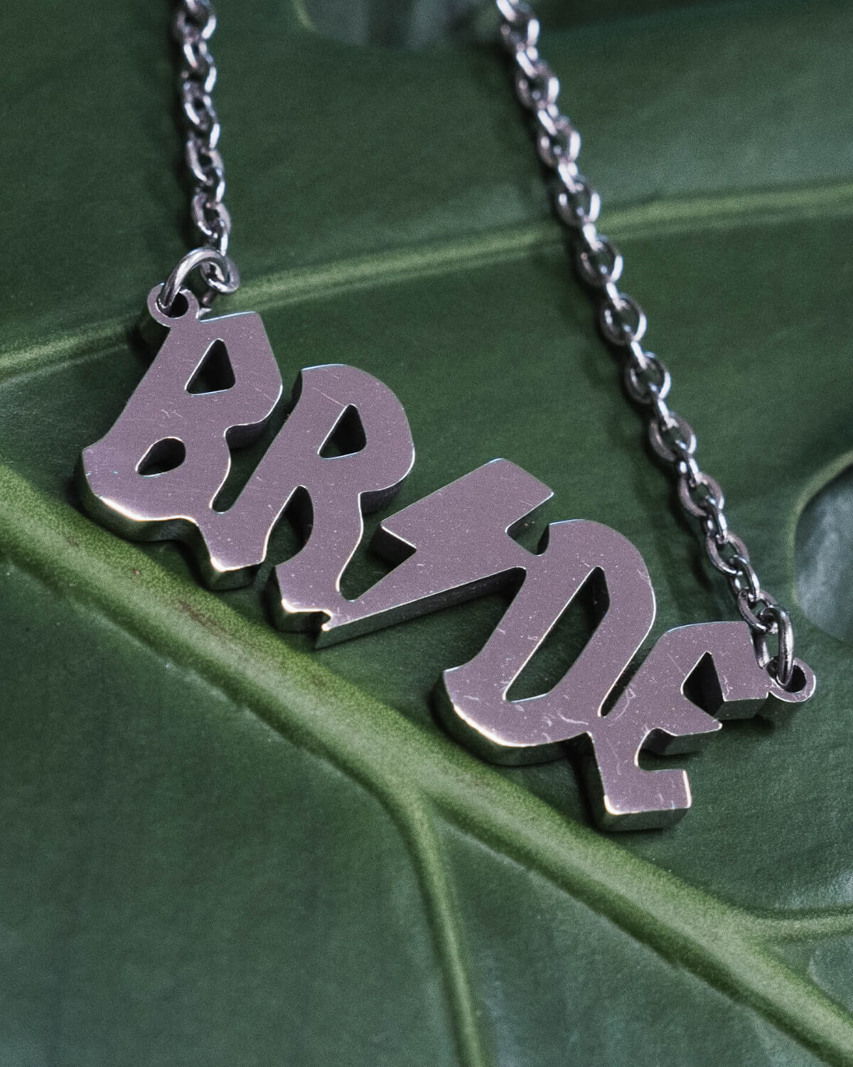 Close-up of a silver “BRIDE” necklace, featuring bold lightning bolt typography, displayed on a green monstera leaf for a dramatic and modern aesthetic.
