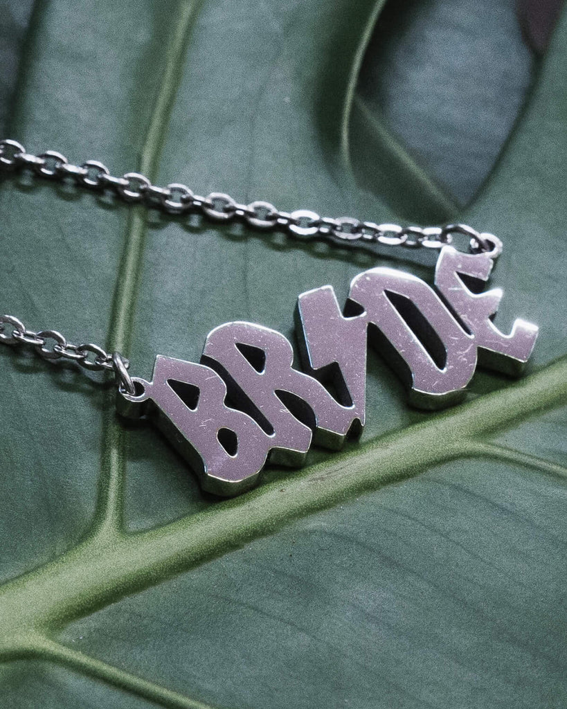 Close-up of a silver “BRIDE” necklace, featuring bold lightning bolt typography, displayed on a green monstera leaf for a dramatic and modern aesthetic.