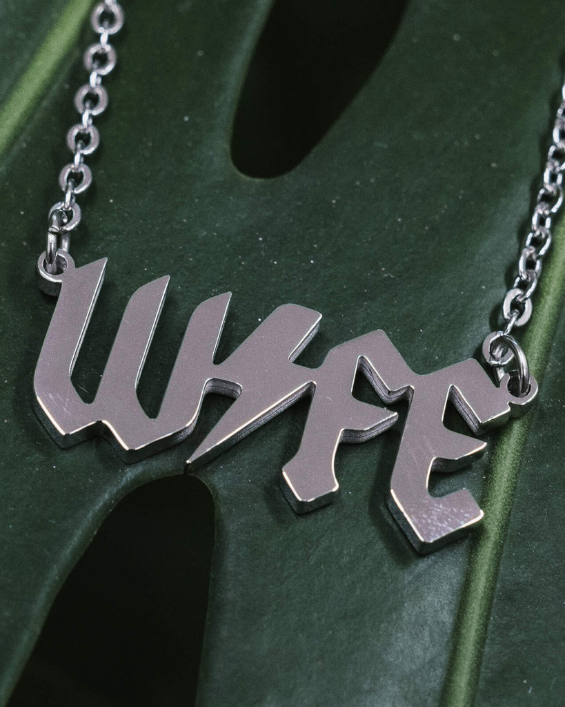 Close-up of a silver metal necklace with the word “WIFE” in a bold, gothic font, featuring a lightning bolt through the middle, displayed on a green monstera leaf background.