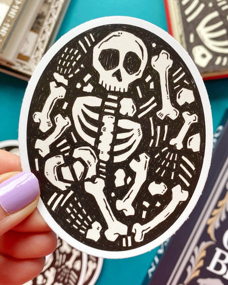 A hand with lilac nail polish holding an oval-shaped matte vinyl sticker featuring a black-and-white illustration of a skeleton with scattered bones on a textured background. A turquoise surface and books are blurred in the background.