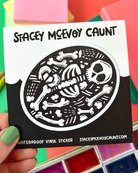  A hand with green nail polish holding the same skeleton sticker, mounted on a branded backing card with “Stacey McEvoy Caunt” written above. Colourful notebooks and art supplies are in the background.

