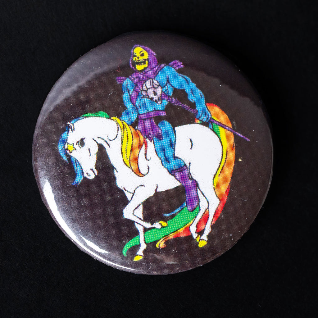 A high-gloss 38mm pin badge featuring Skeletor riding a rainbow-maned unicorn with a black background. A quirky pop culture-inspired design by Bite Your Granny.