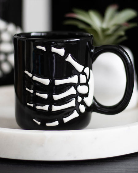 Close-up of a black ceramic mug featuring a raised white skeleton hand design, perfect for gothic or Halloween homeware lovers.