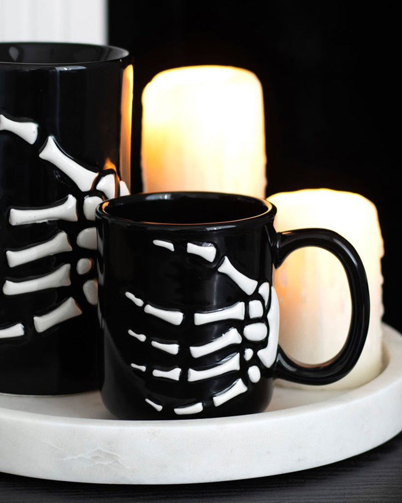 Black ceramic skeleton hand mug on a white tray with glowing candles behind it, creating a spooky Halloween ambience.