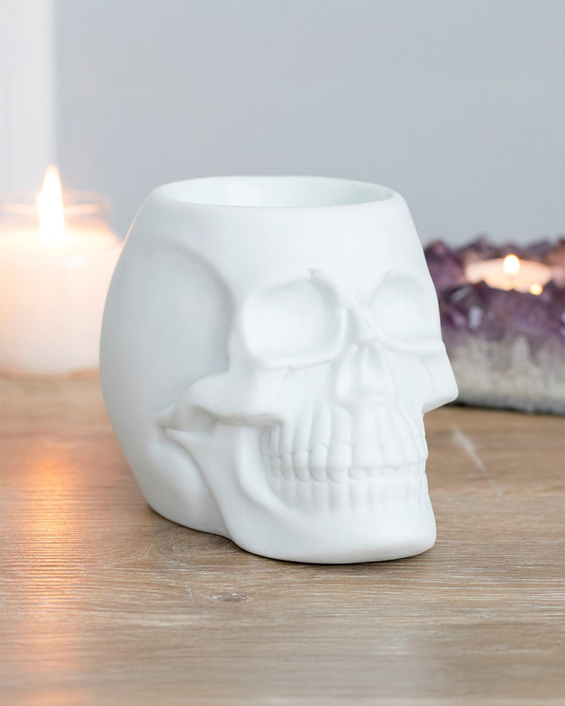 A White Skull Oil Burner by Rebel Romance sits on a wooden surface, surrounded by candles and amethyst crystals. The matte ceramic finish gives it a gothic, modern look, perfect for alternative home decor.