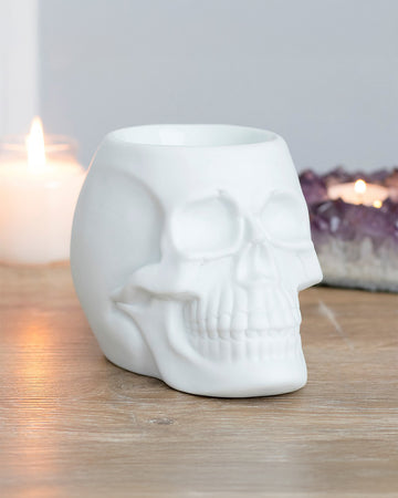 A White Skull Oil Burner by Rebel Romance sits on a wooden surface, surrounded by candles and amethyst crystals. The matte ceramic finish gives it a gothic, modern look, perfect for alternative home decor.