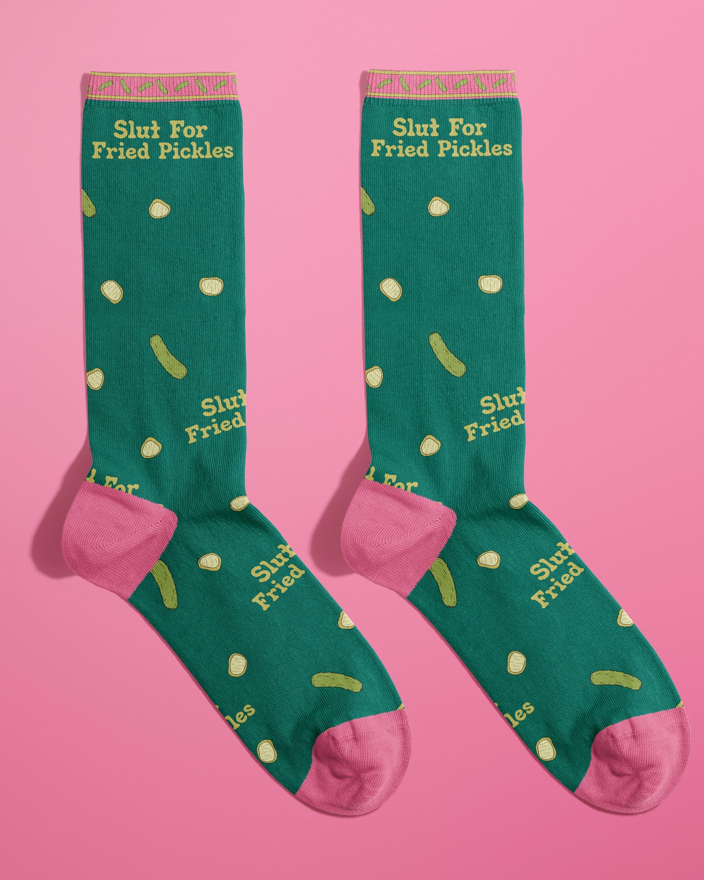 Pair of Rebel Romance’s “Slut For Fried Pickles” crew socks by FUN CLUB displayed side by side on a bright pink background. Featuring green fabric with cartoon pickles, pink toes and heels, and sassy slogan text. A bold and funny fashion gift for foodies and alternative style fans.