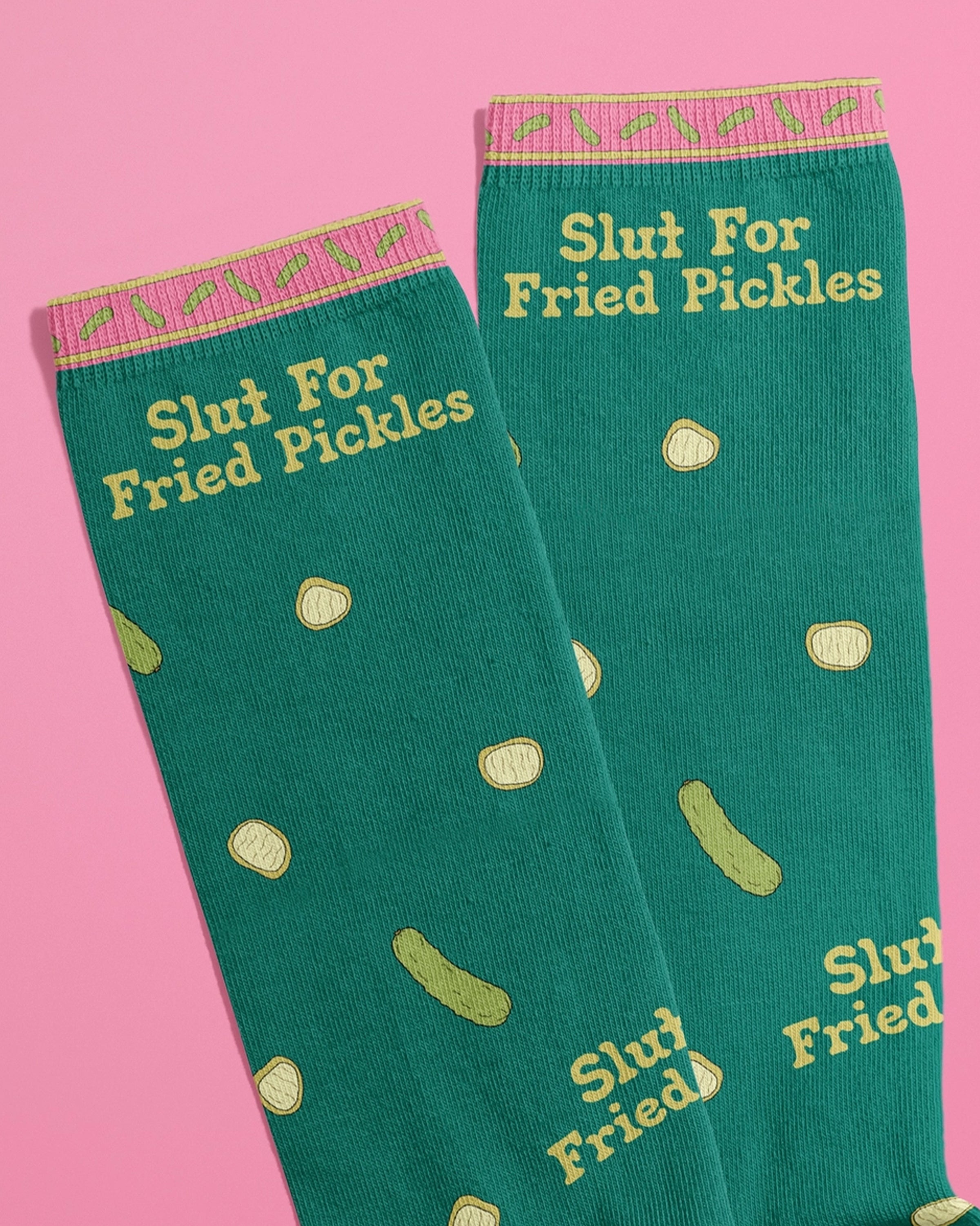 Flat lay of Rebel Romance novelty socks by FUN CLUB with the phrase “Slut For Fried Pickles” in yellow text on green fabric, scattered with pickle slice illustrations. The top band features a pink and yellow pickle pattern. Shot on a pink background. Ideal for alternative fashion lovers and quirky gift seekers.