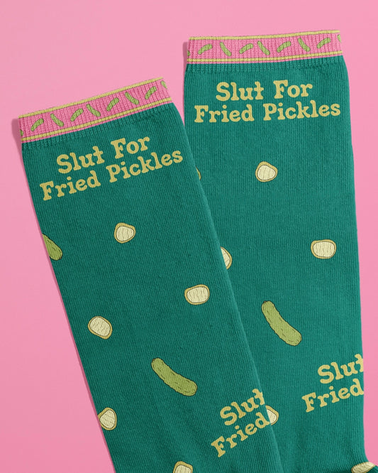 Flat lay of Rebel Romance novelty socks by FUN CLUB with the phrase “Slut For Fried Pickles” in yellow text on green fabric, scattered with pickle slice illustrations. The top band features a pink and yellow pickle pattern. Shot on a pink background. Ideal for alternative fashion lovers and quirky gift seekers.