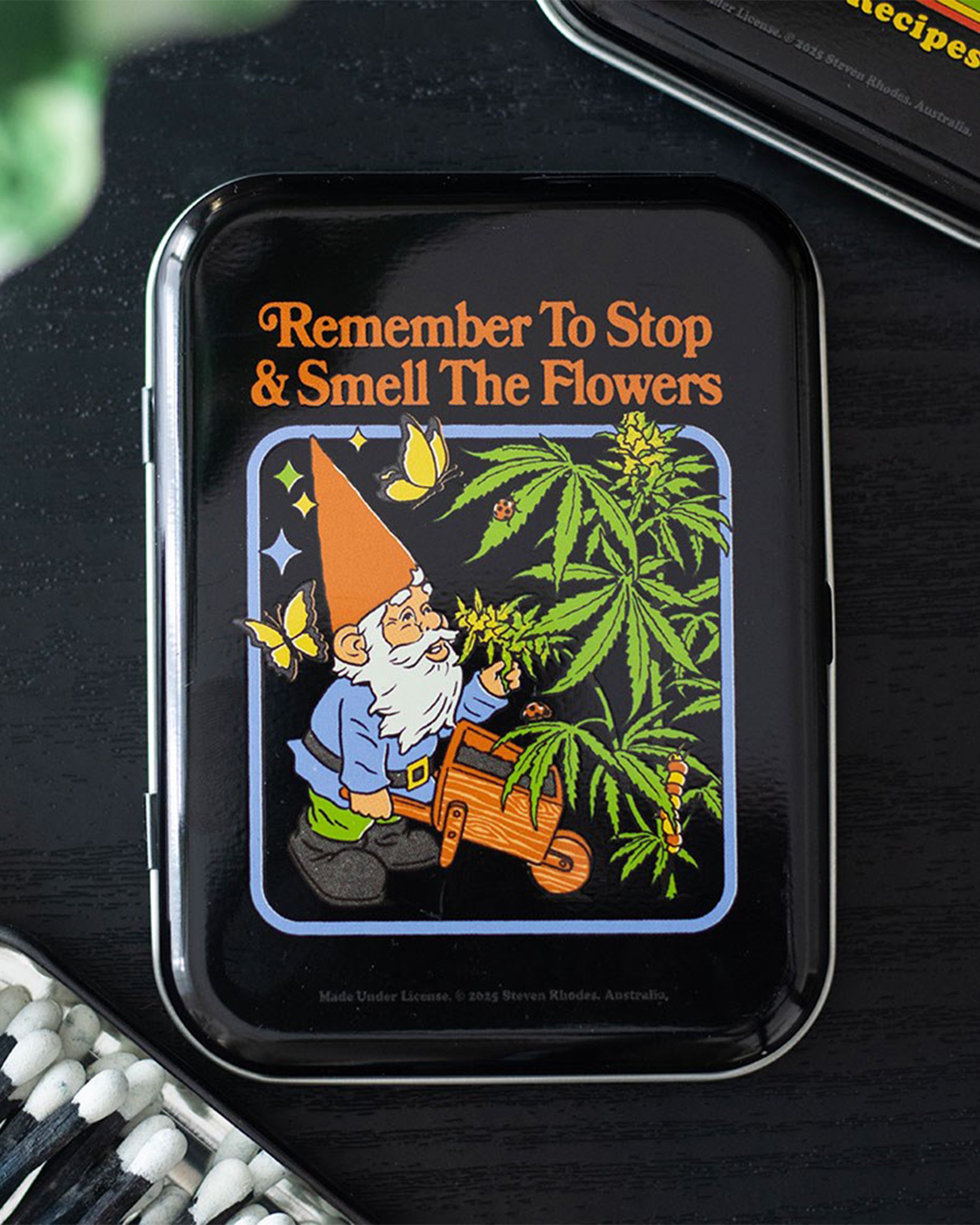 Styled flat lay of the Smell the Flowers stash tin by Steven Rhodes, sold at Rebel Romance. Featuring a retro gnome and cannabis design, this quirky tin is ideal for stashing rolling gear or small keepsakes.