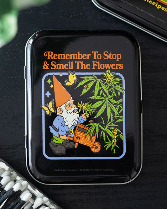 Styled flat lay of the Smell the Flowers stash tin by Steven Rhodes, sold at Rebel Romance. Featuring a retro gnome and cannabis design, this quirky tin is ideal for stashing rolling gear or small keepsakes.