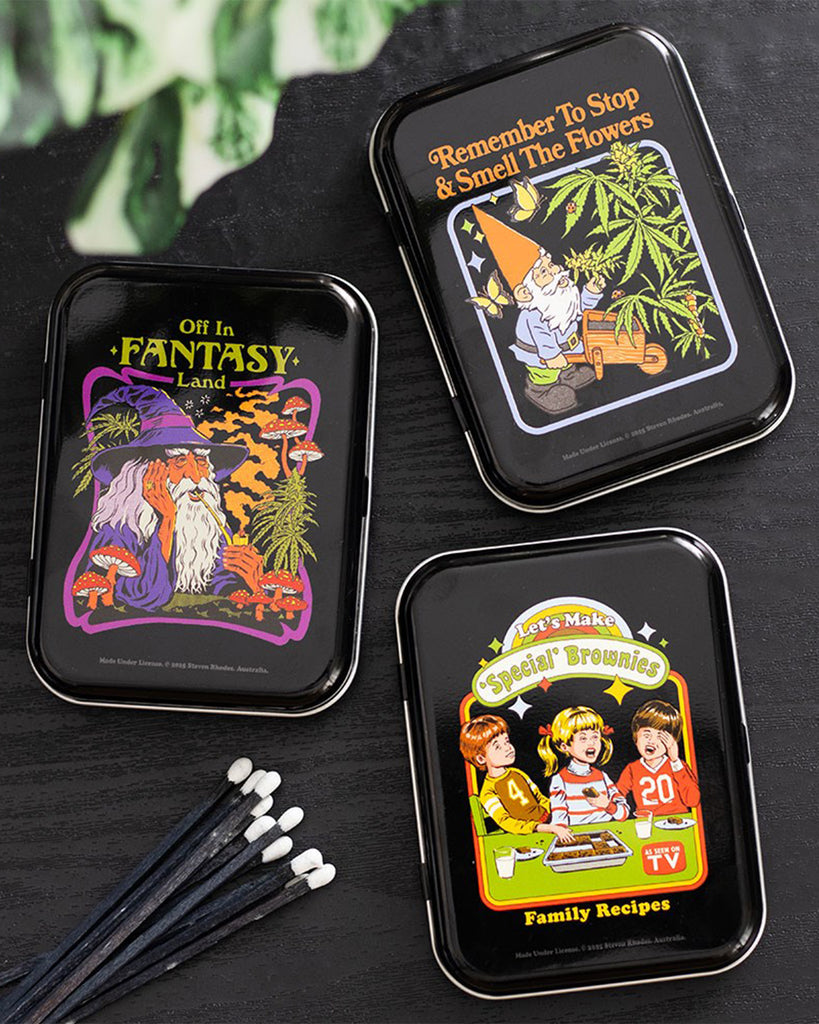 Three Steven Rhodes stash tins sold by Rebel Romance, featuring quirky retro designs. One shows a gnome with cannabis plants and the text "Remember To Stop & Smell The Flowers", another shows a wizard in "Fantasy Land", and the third features kids baking "Special Brownies".