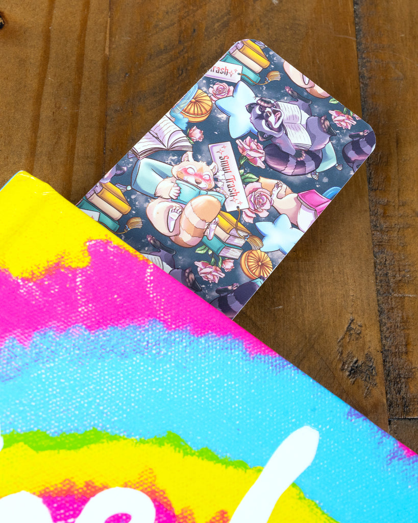  A playful Smut Trash bookmark from Rebel Romance peeking out of a rainbow-toned book, with illustrated animals reading books against a night sky backdrop.