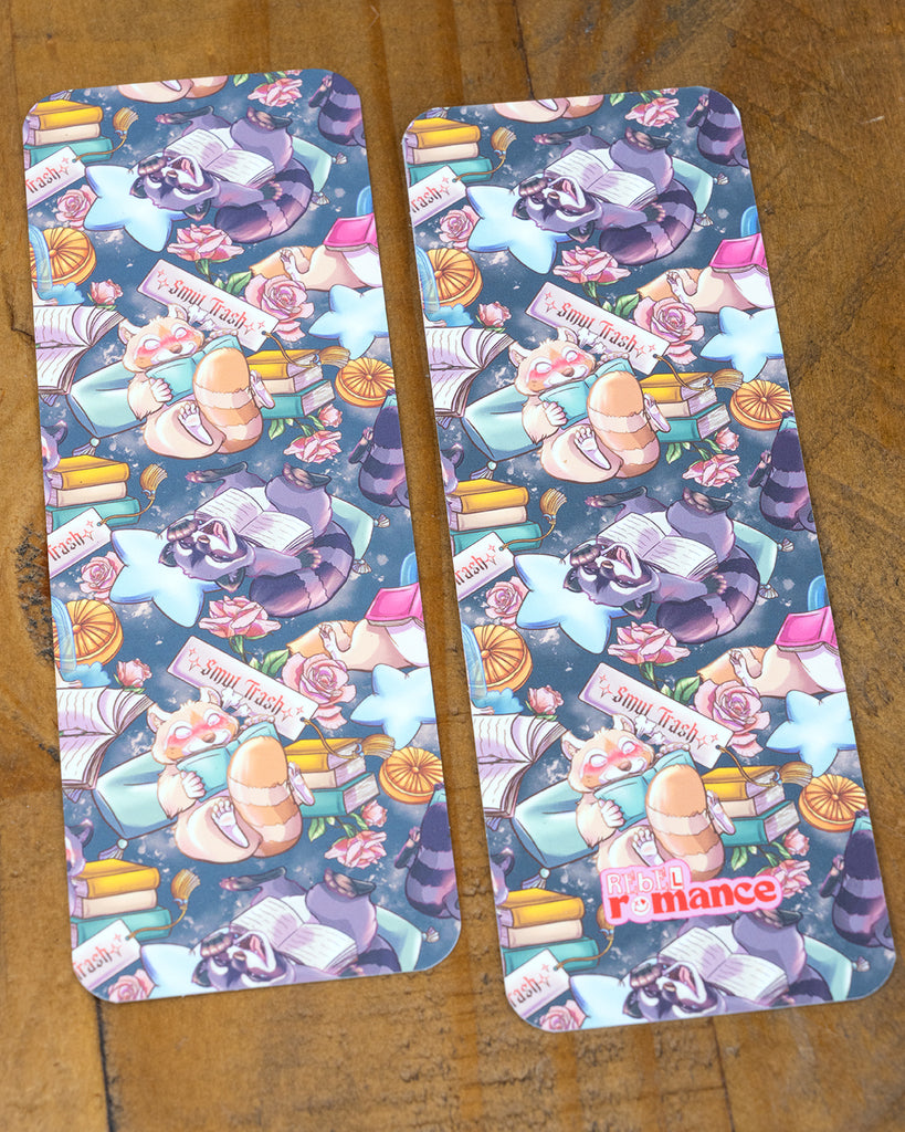 Two Smut Trash bookmarks on a wooden surface, both showing the same whimsical repeat pattern with reading animals, florals, and magical vibes.