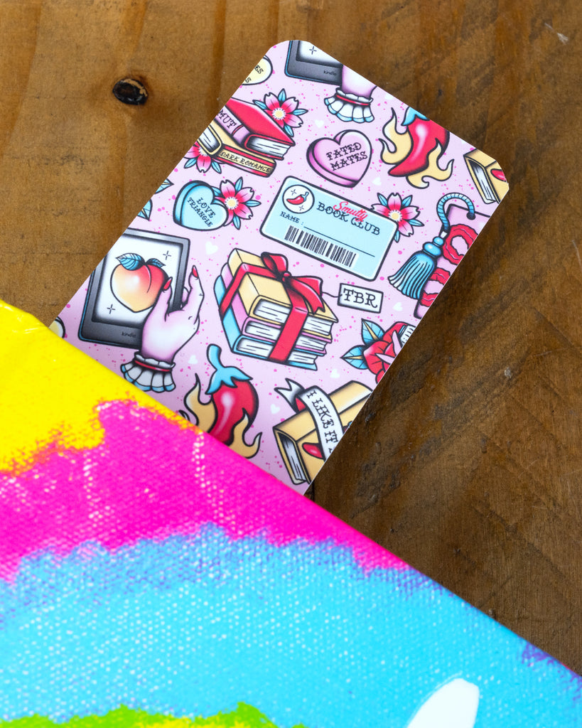 Flatlay of two identical pastel pink Smutty Book Club bookmarks on a wooden background. The bookmarks feature books, hearts, flowers, a barcode name badge, flames, and a hand holding a peach emoji on a phone. Designed by Rebel Romance for romance and spice book lovers.