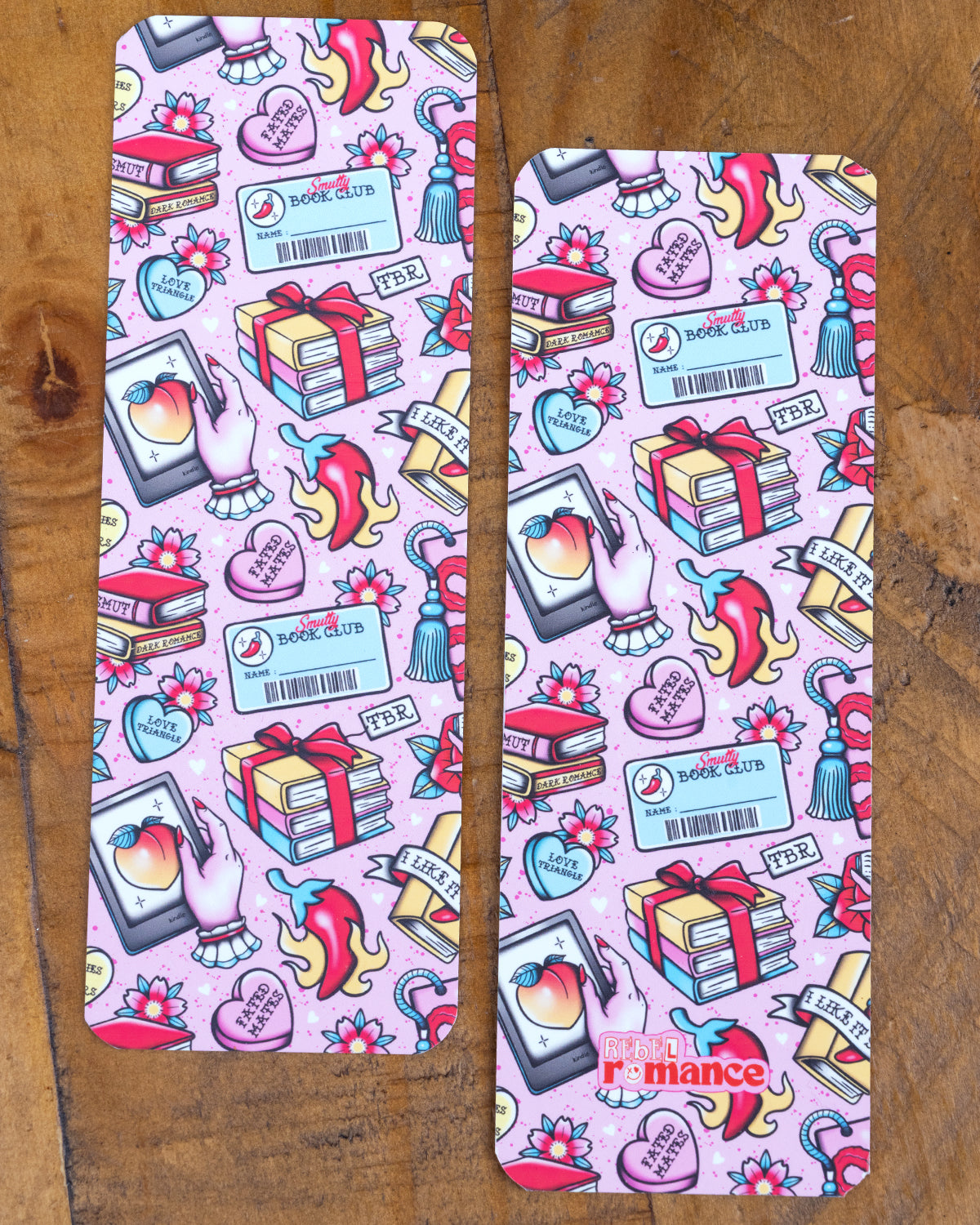 Two pink bookmarks with a smutty romance book club design laid side-by-side on a wooden surface. The design includes hearts, books, peaches, and floral motifs.