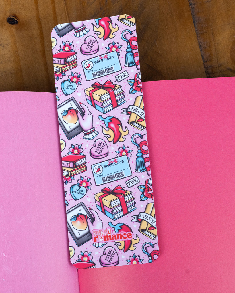 One Smutty Book Club bookmark placed on top of an open pink book, showing off the detailed pink design with books, hearts, flames, and florals.