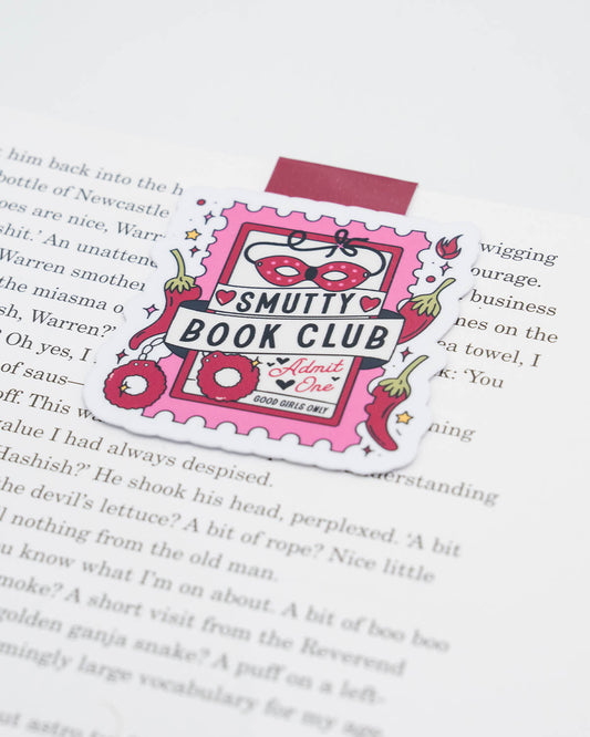 Magnetic bookmark clipped to a book page, designed like a cheeky pink ticket with the text “Smutty Book Club – Admit One.” Decorated with a red mask, fuzzy handcuffs, chilli peppers, and hearts. A playful accessory for romance and spicy book lovers. Perfect for millennials who enjoy alternative humour and bold, flirty stationery. From UK small business Rebel Romance.