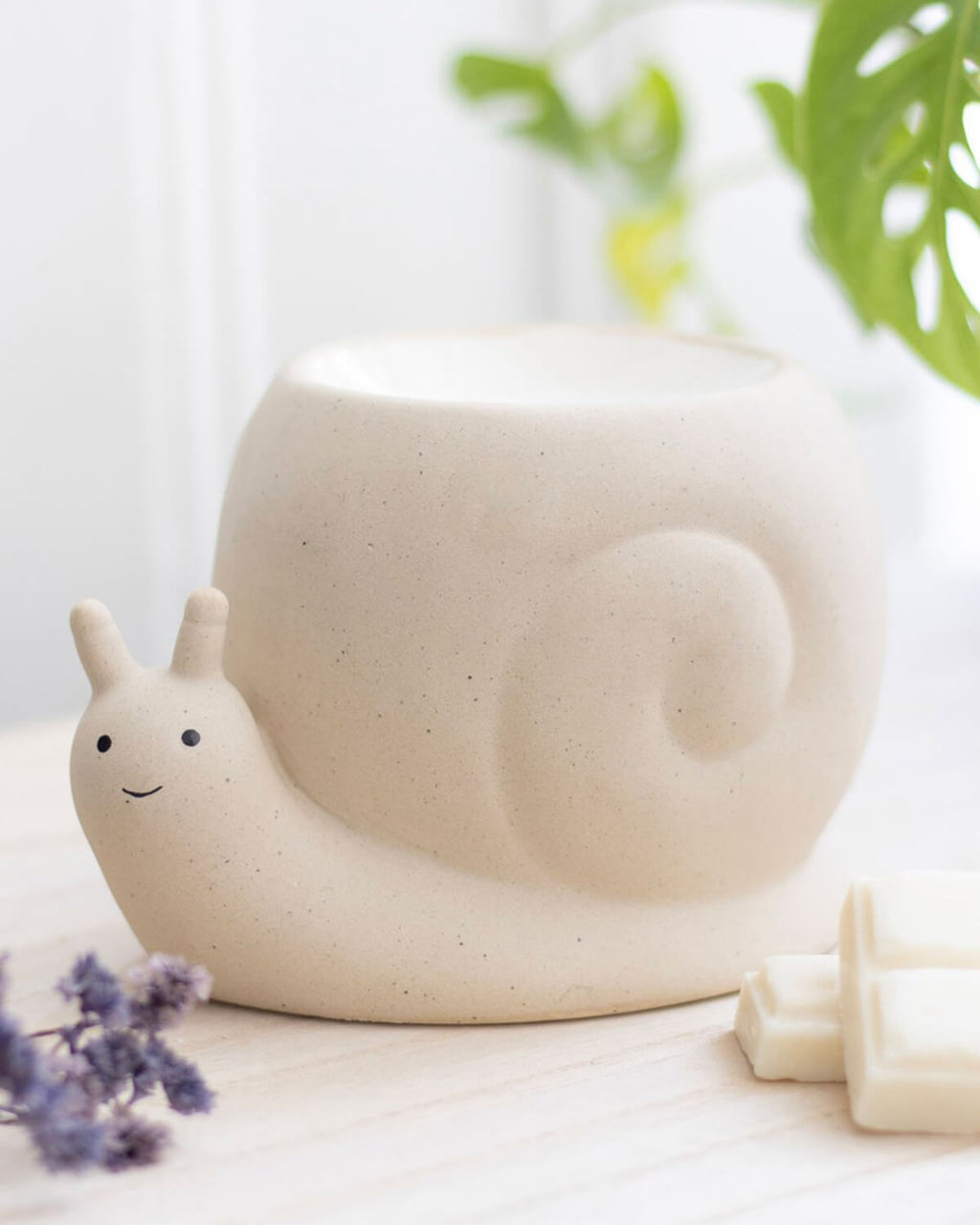 A beautifully styled close-up of a ceramic snail oil burner by Rebel Romance, placed on a wooden surface with wax melts and lavender nearby. The soft, neutral tones and natural lighting highlight its cute, minimalistic design, perfect for creating a cosy and magical home atmosphere. A must-have for fans of quirky and alternative homeware.
