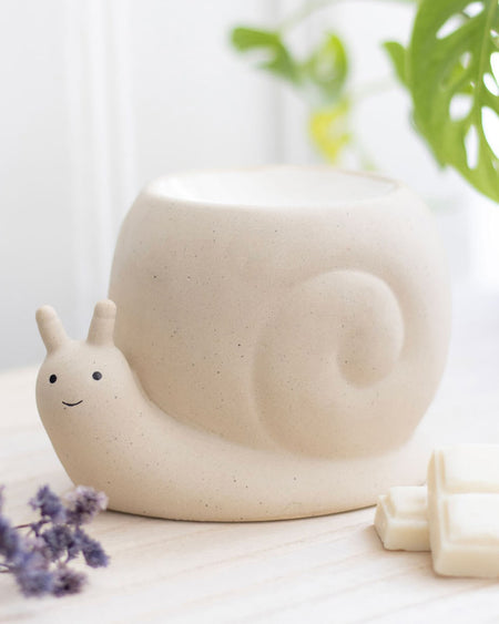A beautifully styled close-up of a ceramic snail oil burner by Rebel Romance, placed on a wooden surface with wax melts and lavender nearby. The soft, neutral tones and natural lighting highlight its cute, minimalistic design, perfect for creating a cosy and magical home atmosphere. A must-have for fans of quirky and alternative homeware.