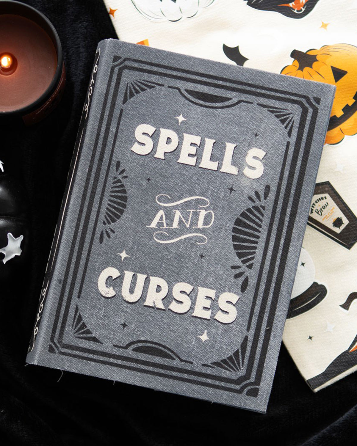 A close-up of the Spells and Curses Book Shaped Storage Box stocked by Rebel Romance, styled with Halloween-themed textiles and candles. The textured grey cover and eerie vintage inspired lettering make it a must-have for a witchy house aesthetic.