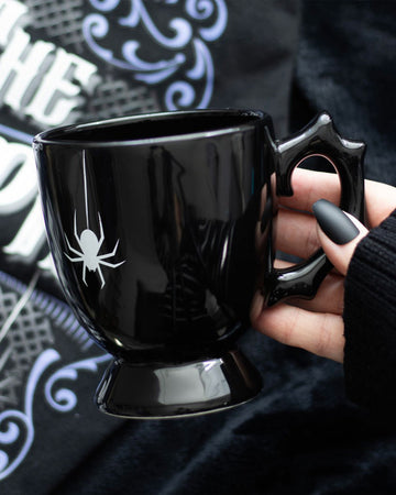 Close-up of a hand holding the Black Spider Teacup with a white spider print and sculpted thorn-like handle.