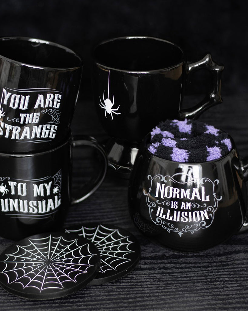 Flat lay of four gothic-style black mugs with spider web coasters and slogan prints including 'Normal is an Illusion' and 'You Are the Strange'.