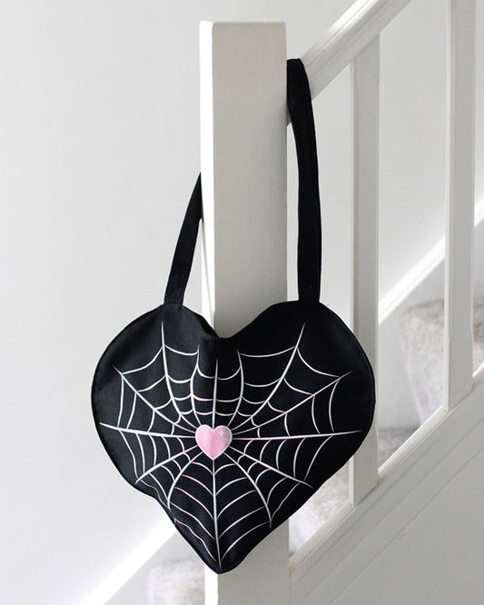Rebel Romance Spiderweb Heart Tote Bag hanging on a white staircase bannister. The black heart-shaped tote features a spiderweb and pink heart design. Perfect for everyday alternative style.