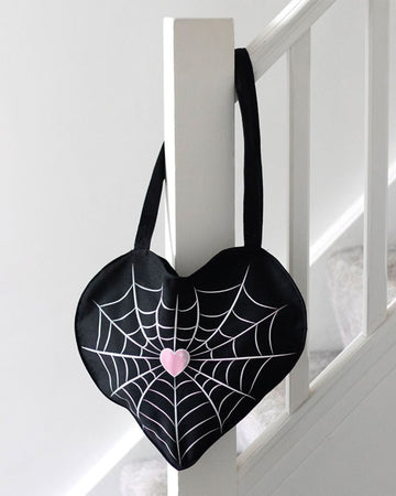 Rebel Romance Spiderweb Heart Tote Bag hanging on a white staircase bannister. The black heart-shaped tote features a spiderweb and pink heart design. Perfect for everyday alternative style.