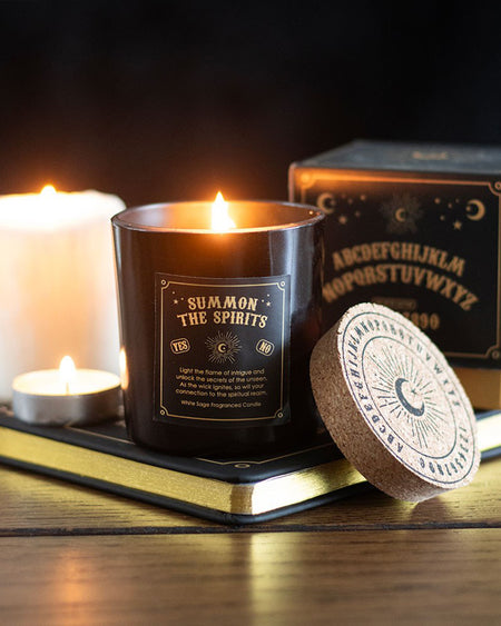 A lit Summon the Spirits white sage candle in a black glass jar with gold detailing, placed on a wooden surface with a book, candles, and a cork lid featuring a celestial design. The scene is atmospheric and mystical.