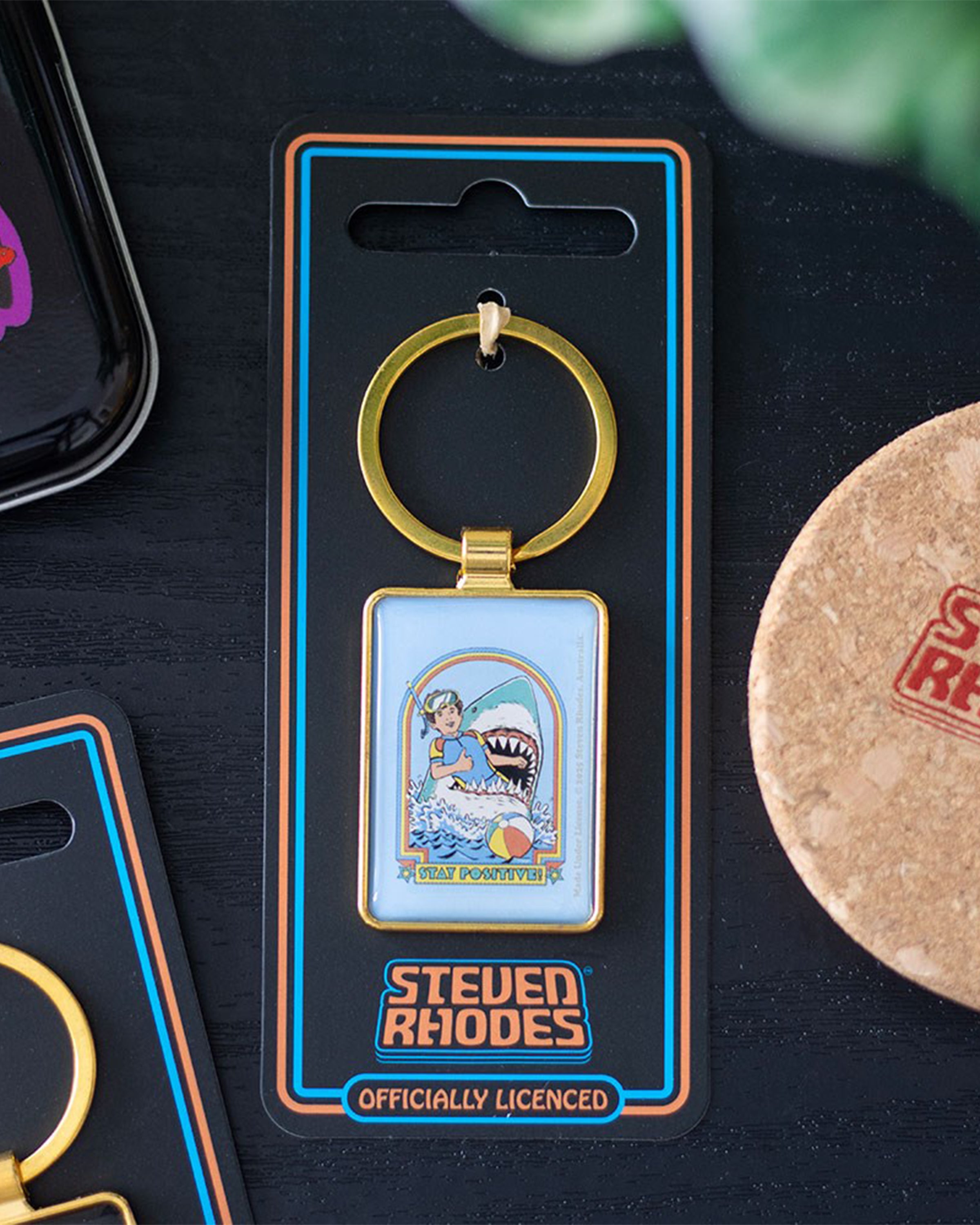 Lifestyle shot of the Rebel Romance Steven Rhodes Stay Positive enamel keyring in packaging on a table. Retro snorkeller and shark design, officially licensed Steven Rhodes accessory. Fun, dark humour gift for alternative millennials.