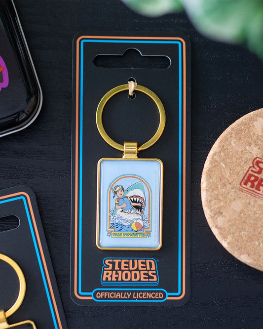 Lifestyle shot of the Rebel Romance Steven Rhodes Stay Positive enamel keyring in packaging on a table. Retro snorkeller and shark design, officially licensed Steven Rhodes accessory. Fun, dark humour gift for alternative millennials.