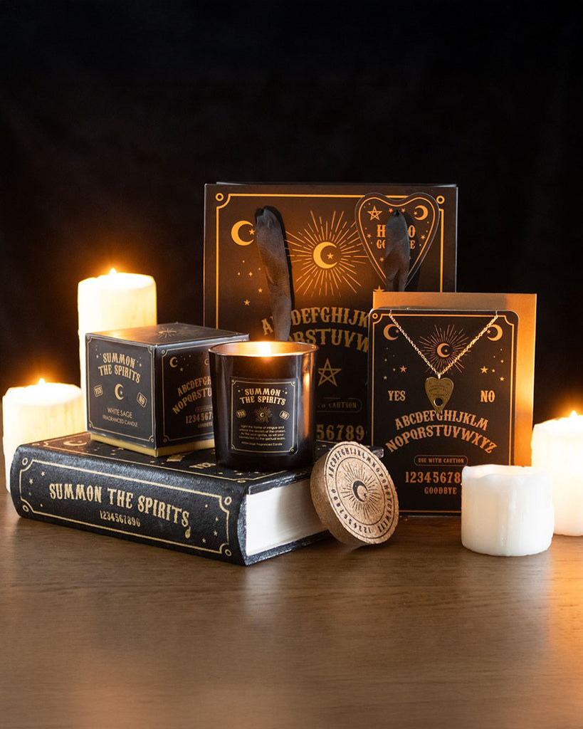 A mystical-themed gift selection from Rebel Romance, featuring the Black Talking Board Book Storage Box with a black and gold Ouija board-inspired design, surrounded by candles, a feather, a candle, and a planchette. The box resembles a book with "Summon the Spirits" on the spine, offering discreet storage for small treasures.