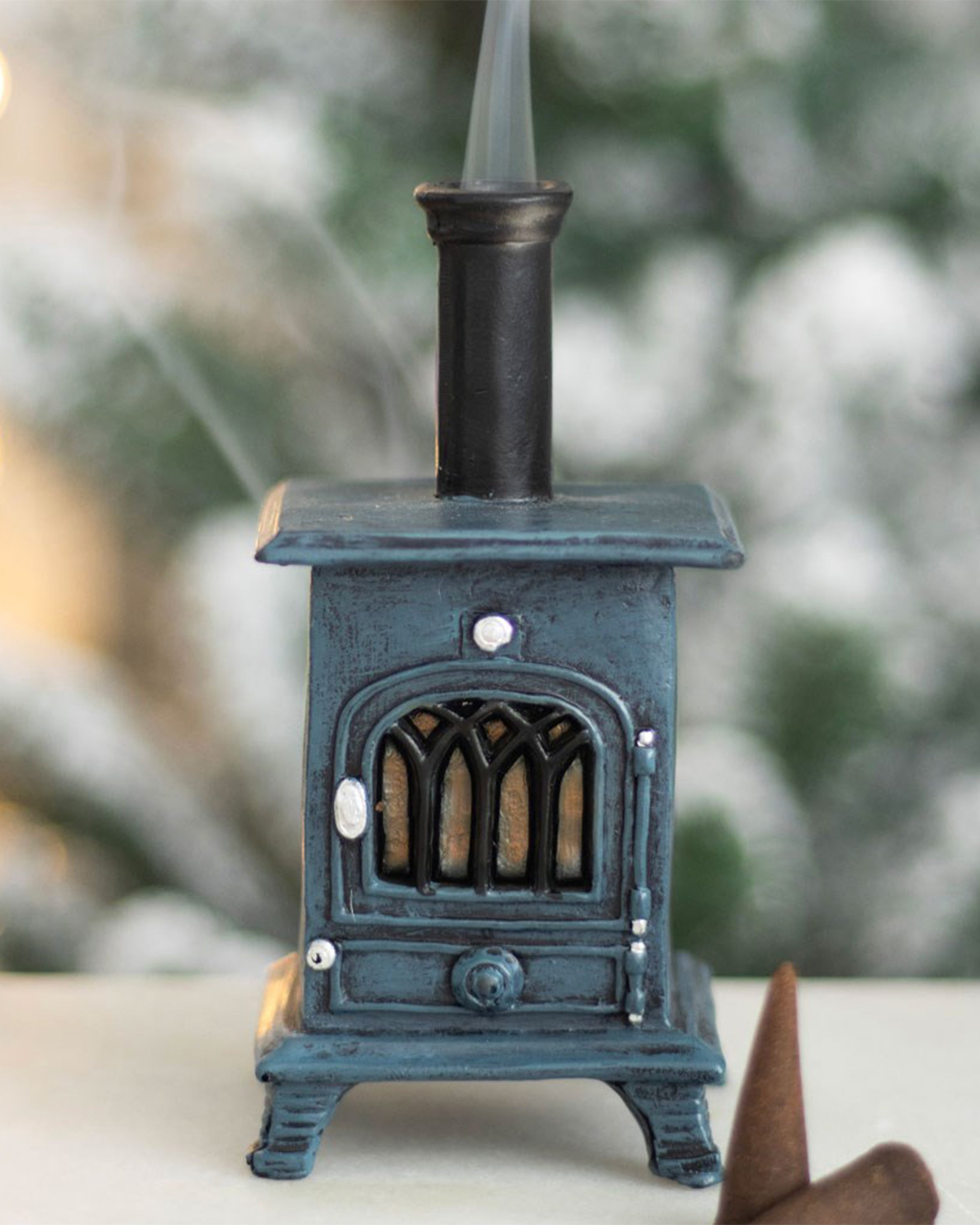 Lifestyle image of the Rebel Romance Stove Incense Cone Burner with incense cones and a blurred festive background. Smoke swirls from the chimney for a magical winter vibe.