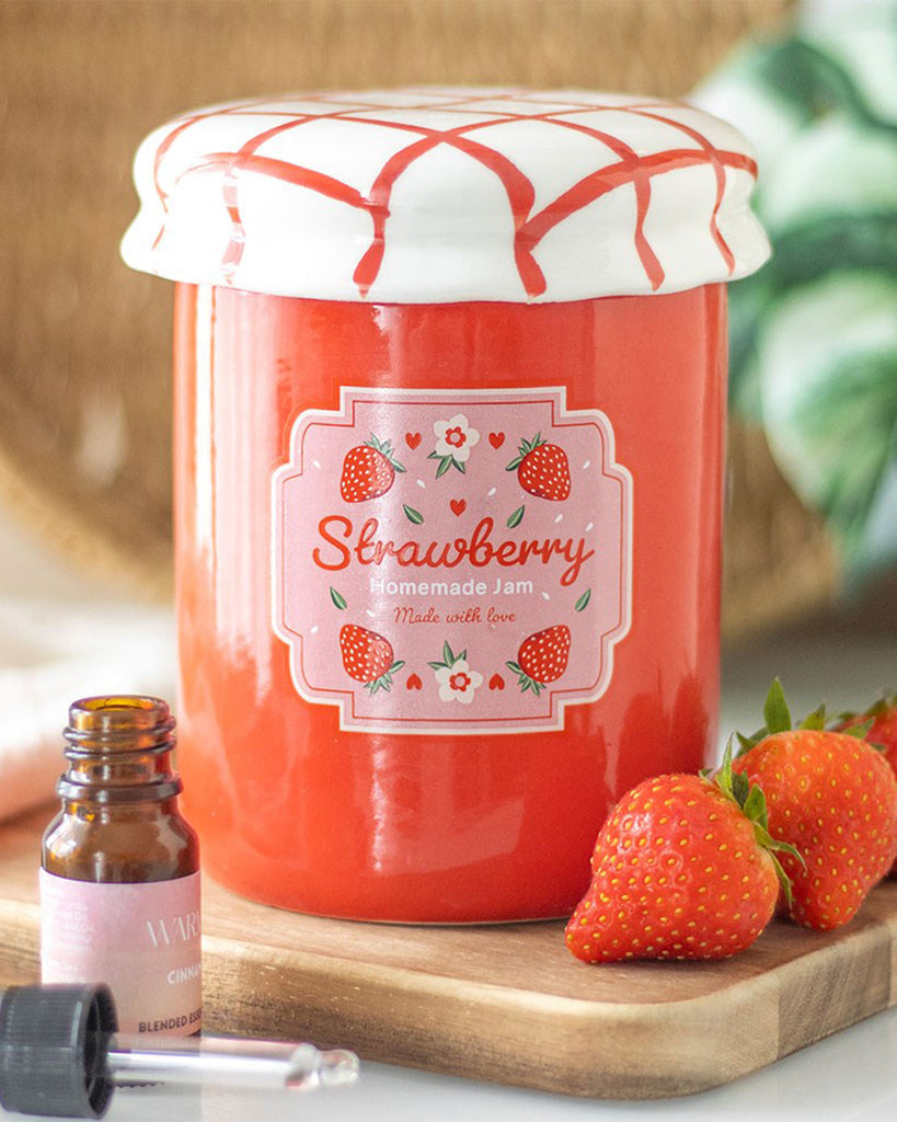 Cosy home decor featuring a strawberry jam jar oil burner: The ceramic oil burner sits on a wooden tray, surrounded by fresh strawberries and a cinnamon-scented essential oil bottle. The lid is closed, making it look like a delightful decor piece.