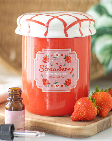 Cosy home decor featuring a strawberry jam jar oil burner: The ceramic oil burner sits on a wooden tray, surrounded by fresh strawberries and a cinnamon-scented essential oil bottle. The lid is closed, making it look like a delightful decor piece.