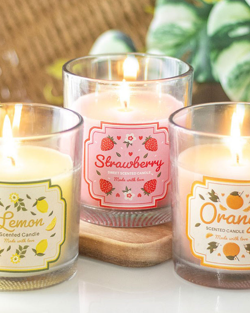 Set of three scented fruit candles from Rebel Romance in Lemon, Strawberry, and Orange varieties, lit and styled together in glass jars with cute illustrated labels. Fruity home décor from an ethical UK small business.