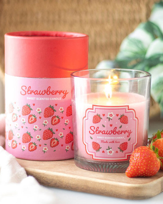 Glass Strawberry Scented Candle by Rebel Romance lit beside its matching pink and red illustrated box, displayed with fresh strawberries on a wooden board. A fun fruity home décor gift from a UK small business.