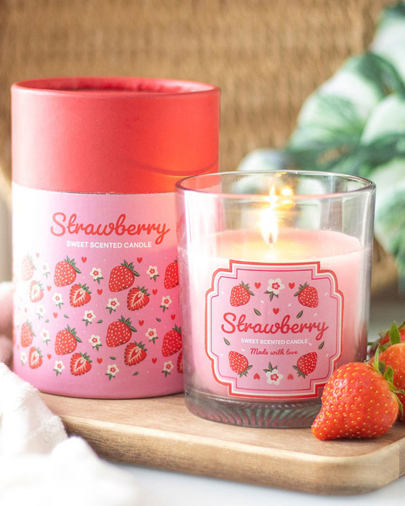 Glass Strawberry Scented Candle by Rebel Romance lit beside its matching pink and red illustrated box, displayed with fresh strawberries on a wooden board. A fun fruity home décor gift from a UK small business.