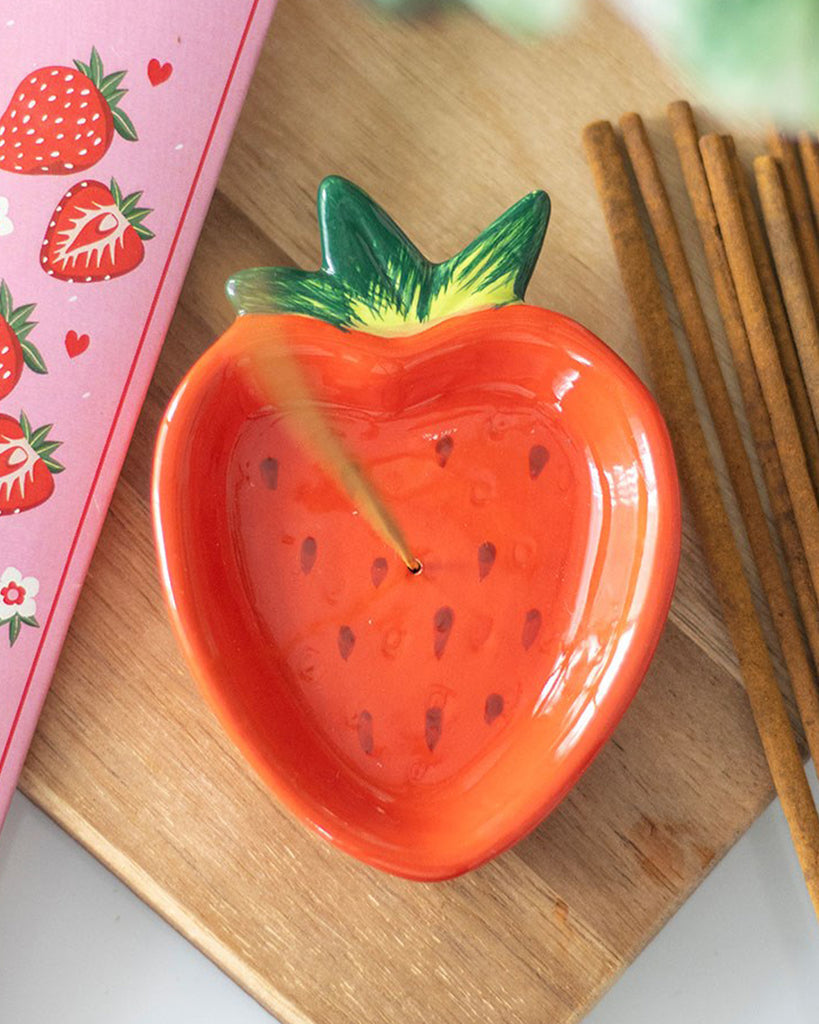 A styled shot of the strawberry incense holder on a wooden surface, surrounded by incense sticks and a strawberry-themed notebook. A perfect gift for fruity and retro décor lovers.