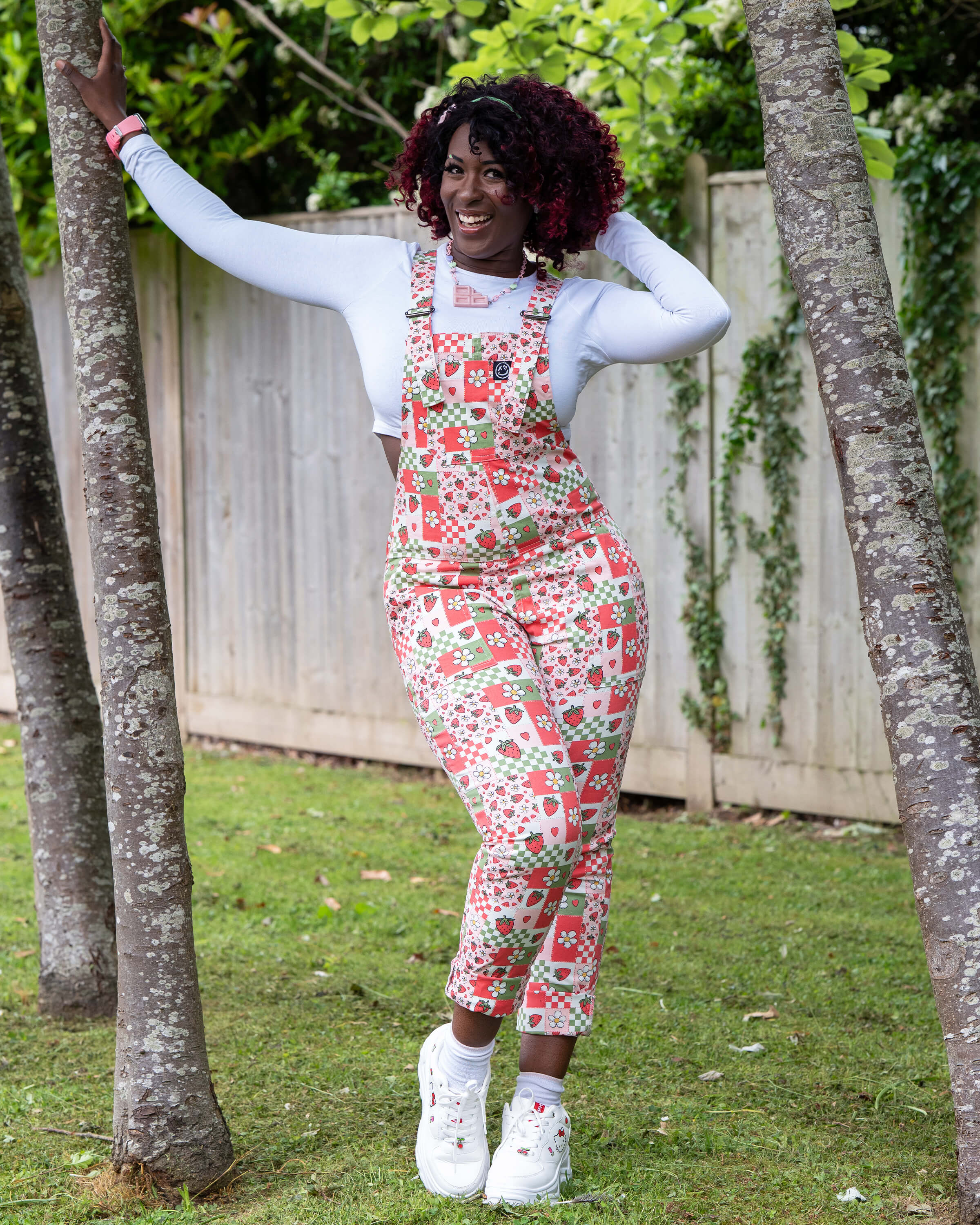 Ayesha poses outdoors in the Rebel Romance Strawberry Picnic Stretch Dungarees, leaning between two trees. The fitted dungarees feature a red, pink, and green patchwork print with strawberries, daisies, hearts, and checks. Styled with a long sleeve white top and chunky trainers.
