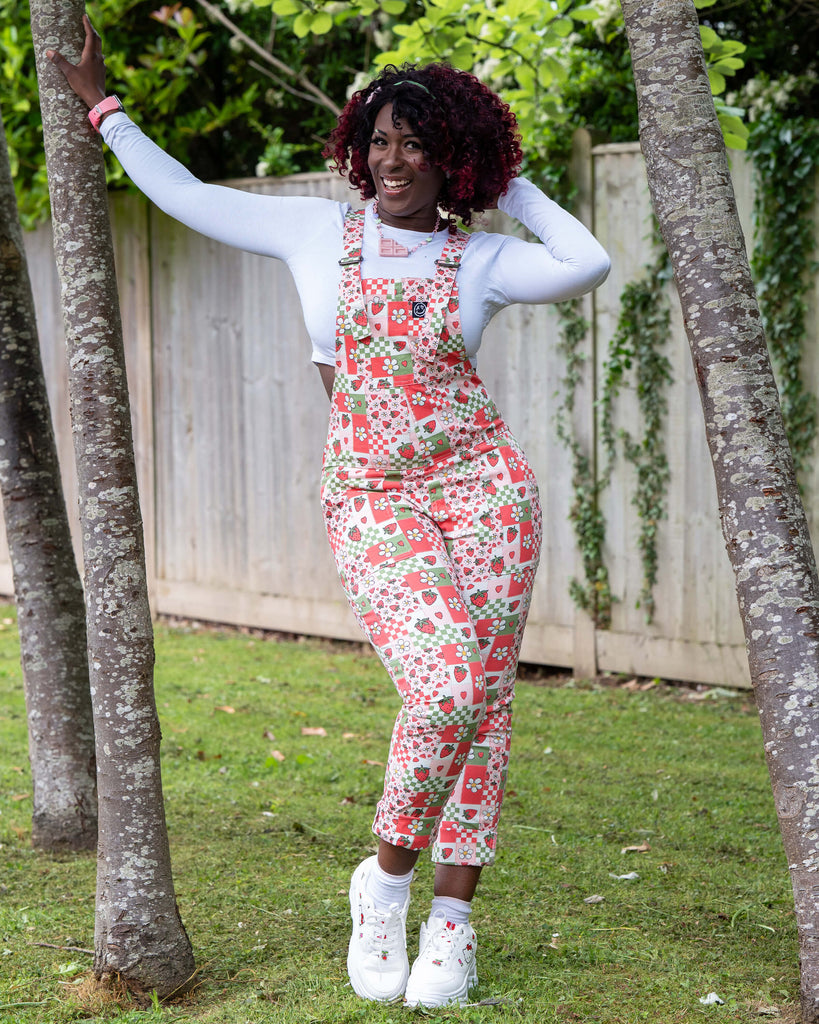 Ayesha poses outdoors in the Rebel Romance Strawberry Picnic Stretch Dungarees, leaning between two trees. The fitted dungarees feature a red, pink, and green patchwork print with strawberries, daisies, hearts, and checks. Styled with a long sleeve white top and chunky trainers.