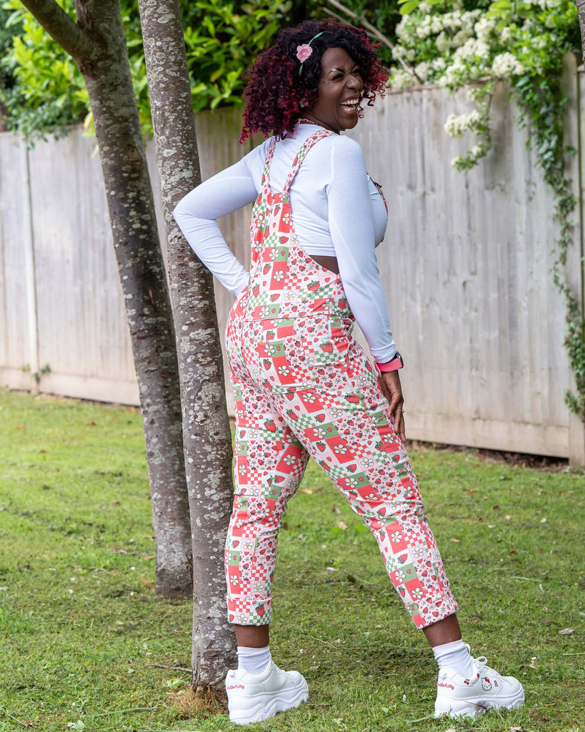 Rear view of Ayesha wearing the Rebel Romance Strawberry Picnic Stretch Dungarees, showing the vibrant back detail and pockets. The dungarees are covered in strawberries, flowers, and hearts. Styled with a white top and platform trainers.