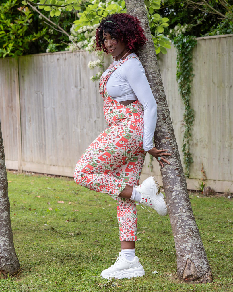 Side view of Ayesha modelling the Rebel Romance Strawberry Picnic Stretch Dungarees, leaning against a tree. The vibrant dungarees feature strawberries, daisies, hearts, and checks. Styled with a long sleeve white top and white trainers.