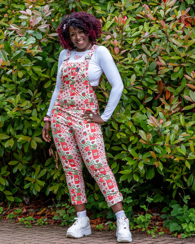 Ayesha smiles in front of a lush green hedge while modelling the Rebel Romance Strawberry Picnic Stretch Dungarees. The bold print includes strawberries, flowers, and hearts in a pink, red, and green colour palette. Styled with white platform trainers.
