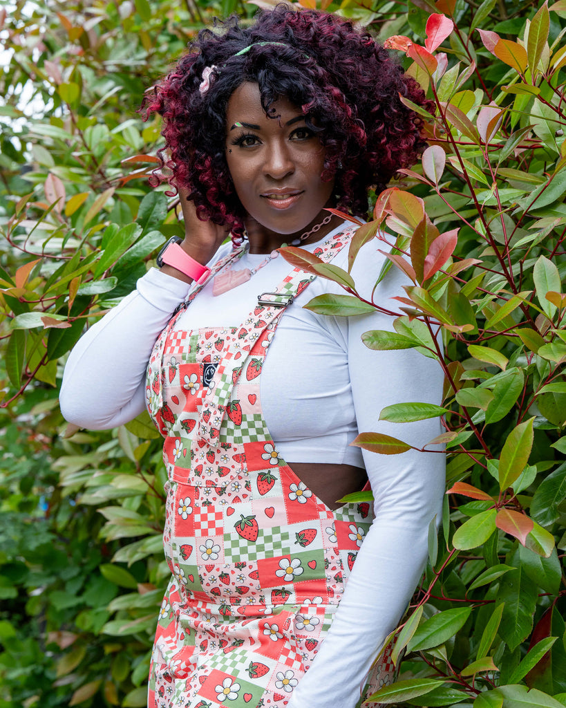 Close-up portrait of Ayesha in the Rebel Romance Strawberry Picnic Stretch Dungarees, surrounded by leafy greenery. The print includes strawberries, daisies, and checkerboard patches. Styled with a white top and pink accessories.