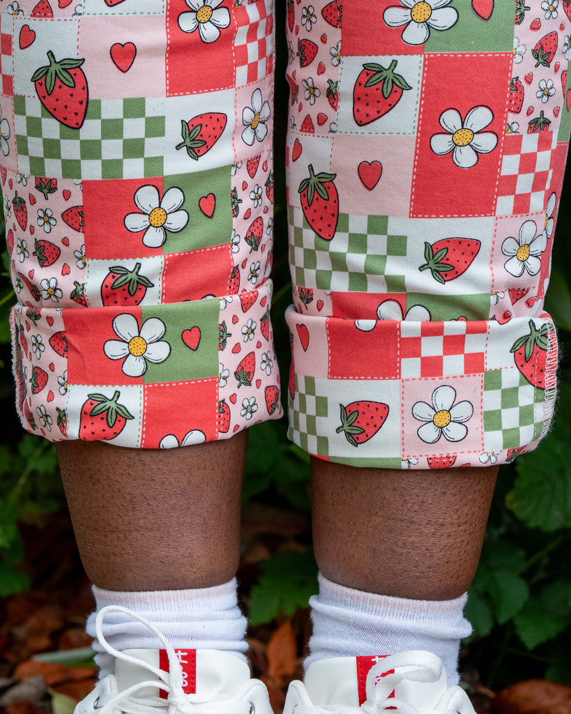 Close-up of the rolled cuffs on the Rebel Romance Strawberry Picnic Stretch Dungarees, showing the fun strawberry, daisy, and heart print on red, green, and pink patchwork fabric. Styled with white socks and chunky trainers.