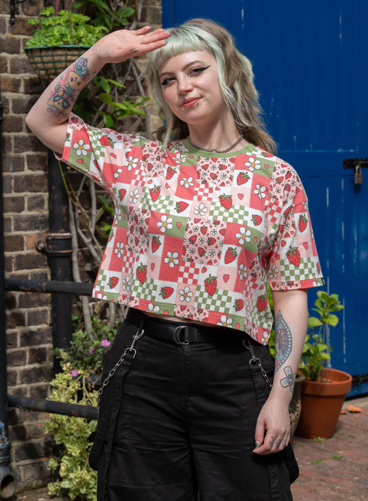 Model wearing a Rebel Romance Strawberry Picnic Cropped Boxy Tee with a retro patchwork print of strawberries, daisies, and checkerboards. The short-sleeve top features a contrast green neckline and relaxed fit.