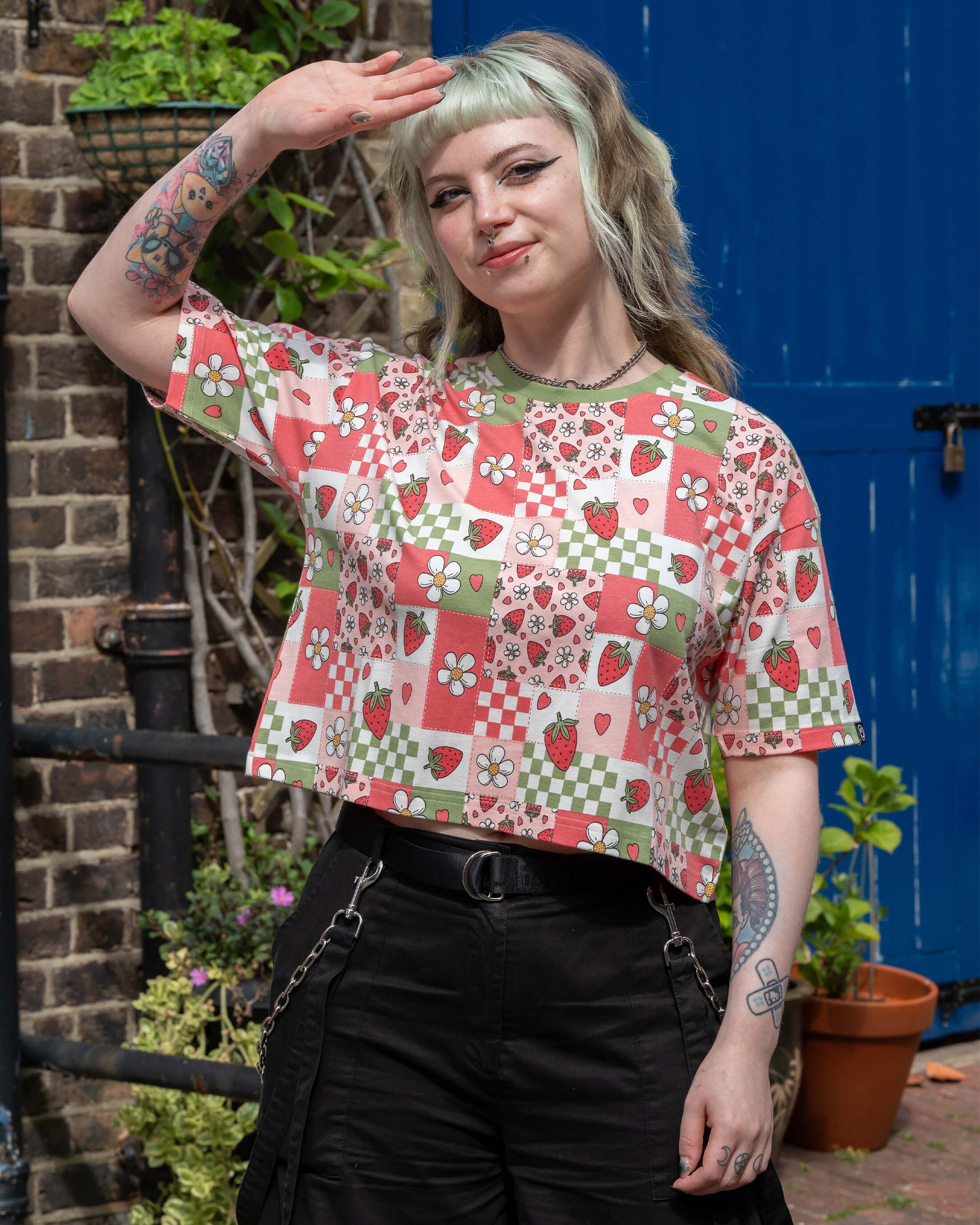 Model wearing a Rebel Romance Strawberry Picnic Cropped Boxy Tee with a retro patchwork print of strawberries, daisies, and checkerboards. The short-sleeve top features a contrast green neckline and relaxed fit.