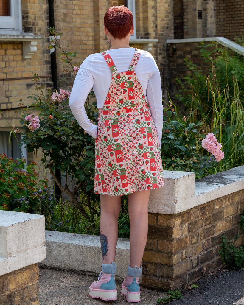 Alternative femme model with tattoos and short purple red hair wearing a strawberry picnic checkerboard patterned dress and pink boot shoes standing outdoors with plants and a building in the background. She has paired it with a white long sleeve underneath and is facing away to show the back detailing. 